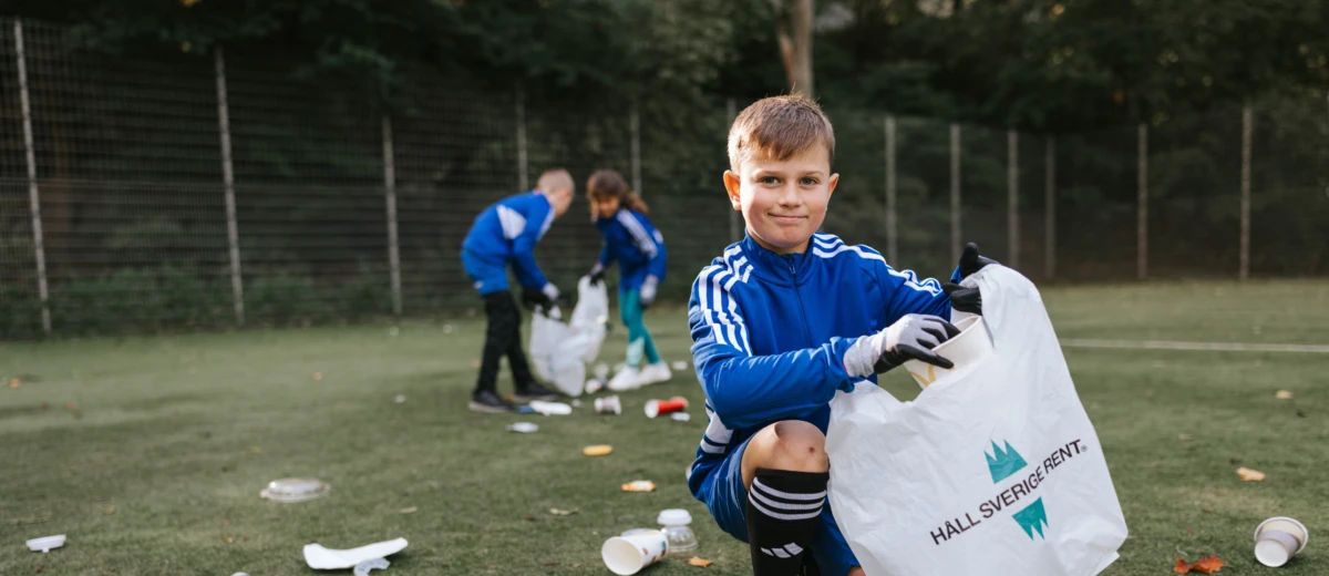 Pojke i fotbollskläder tittar in i kameran medan han plockar skräp